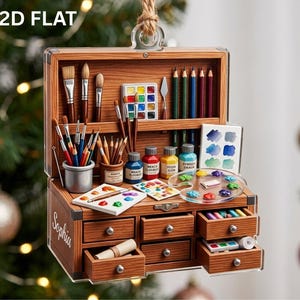 May include: A 2D flat wooden art supply chest ornament featuring the name Sophia on the side. The open chest displays various paintbrushes, colored pencils, a watercolor palette, paint bottles, and small drawers filled with art supplies.