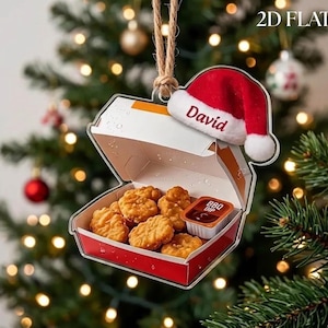 May include: A 2D flat Christmas tree ornament shaped like an open red and white cardboard box filled with golden-brown chicken nuggets and a small container of BBQ dip. A red Santa hat with the name David printed on it sits on the box.