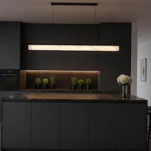 May include: A long, rectangular linear pendant light fixture with a white, marbled, translucent shade suspended by two thin black wires from a black ceiling mount, emitting a soft, warm, diffused glow over a dark kitchen island.