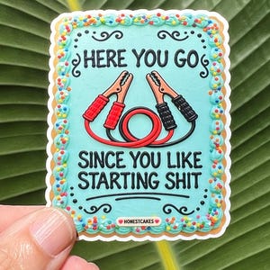May include: A rectangular sticker designed to look like a cake with light blue frosting, colorful sprinkles, and a border of piped icing. It features red and black jumper cables and the text: HERE YOU GO SINCE YOU LIKE STARTING SHIT.