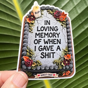 May include: A rectangular sticker shaped like a cake with gray cracked icing, black piped borders, and colorful floral decorations. It features a small candle labeled RIP and the text IN LOVING MEMORY OF WHEN I GAVE A SHIT, with HONEST CAKES at the base.