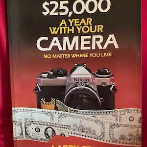 May include: Book cover with a red background and the title "How You Can Make $25,000 A Year With Your Camera" in yellow text. The subtitle is "No Matter Where You Live" in white text. A Nikon camera with a roll of film is in front of the title. The book cover also features a stack of $100 bills and the author's name, "Larry Cribb" in yellow text.