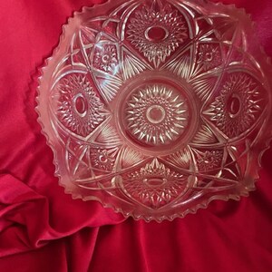 May include: Clear glass serving bowl with a detailed starburst and geometric pattern. The bowl has a scalloped edge and is set against a red background. The bowl's design features repeating oval and star shapes.