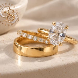 Può includere: Un set di tre anelli in oro composto da un solitario centrale con diamante taglio ovale, una fede impreziosita da diamanti taglio baguette e rotondo, e una fede in oro lucido. Il logo Celeste Siren è visibile nell'angolo in alto a sinistra.
