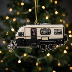 May include: A flat, two-dimensional camper ornament featuring a cream and black exterior with the text THE NOMADIC NOEL printed on the side. The camper has two black wheels, a hitch, and a gold string attached to the top for hanging purposes.