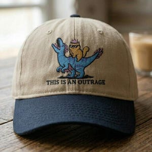 Pode incluir: Um boné de beisebol bege com aba azul-marinho escuro, apresentando um bordado de um dinossauro azul com um gato amarelo usando óculos de sol e uma coroa nas costas, acompanhado pelo texto THIS IS AN OUTRAGE em letras pretas.