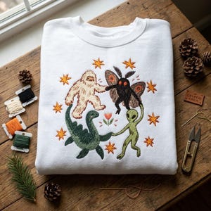 May include: A white crewneck sweatshirt featuring an embroidered design of a beige Bigfoot, a black moth-like creature, a green aquatic dinosaur, and a green alien holding hands in a circle, surrounded by orange stars and a small central flower.