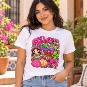 May include: A white t-shirt featuring a colorful graphic print with the text Dulce vida mexicana. The design includes a small doll, a brown clay pot, a cactus, pink and tan concha bread, a pink bow, and a lollipop surrounded by bright flowers.