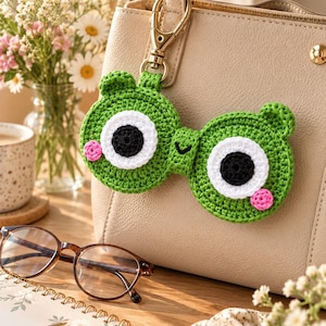 Könnte beinhalten: Ein grüner, gehäkelter Brillenhalter in Froschform mit großen weißen und schwarzen Augen, einem kleinen schwarzen gestickten Mund und rosa kreisförmigen Wangen. Das Accessoire ist mit einem goldenen Karabinerhaken an einer beigen Handtasche befestigt.