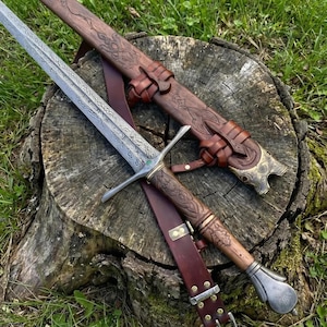 May include: A steel sword with a patterned blade, a brown leather-wrapped handle, and a metal pommel. It is accompanied by a matching brown leather scabbard featuring intricate embossed designs, brass accents, and a leather belt attachment.