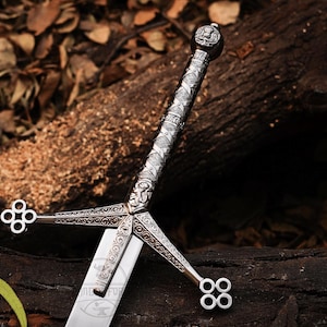 May include: A silver-coloured sword featuring an intricate metal hilt with detailed engravings and a crossguard ending in four-circle clover-like motifs. The handle has a textured, patterned grip and a rounded pommel with a thistle emblem design.