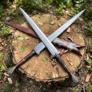 Könnte beinhalten: Zwei zweischneidige Schwerter mit gemusterten Stahlklingen und braunen, lederumwickelten Griffen mit rautenförmigen Knäufen. Die Schwerter werden von einer dunkelbraunen Holzscheide mit metallischen Akzenten und Lederriemen begleitet.