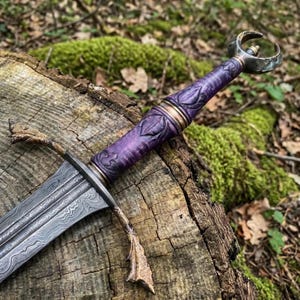 May include: A dagger with a patterned Damascus steel blade, a carved purple wooden handle with bronze-coloured accents, and a crescent-shaped pommel. The crossguard features textured, branch-like bronze-coloured metal detailing.