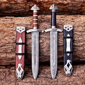 May include: Two daggers with Damascus steel blades and matching scabbards. One dagger has a brown wooden handle and scabbard, while the other has a black handle and scabbard. Both feature silver-coloured metal crossguards, pommels, and eagle motifs.