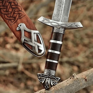 May include: A Damascus steel dagger with a patterned blade and a dark wood handle featuring silver bands. The matching wooden scabbard displays an intricate carved knotwork pattern and a silver metal fitting shaped like a bird of prey.