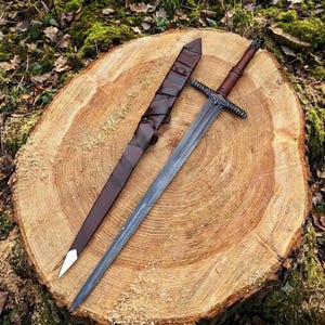 May include: A steel sword with a dark brown leather-wrapped handle and a matching dark brown leather scabbard. The sword features a silver-toned crossguard with intricate etched detailing and a pointed blade, resting alongside its protective sheath.