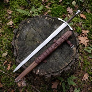 May include: A steel longsword with a dark brown leather-wrapped grip and a matching brown leather scabbard featuring decorative leather lacing, a metal pommel, and a simple crossguard, resting diagonally across a weathered tree stump.