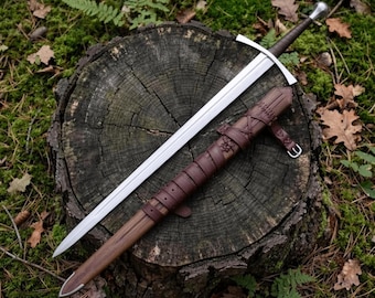 Puede incluir: Una espada larga de acero con empuñadura forrada en cuero marrón oscuro y una vaina de cuero marrón a juego con cordones decorativos, un pomo de metal y una guarda sencilla, colocada en diagonal sobre un tocón de árbol.