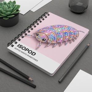 May include: A spiral-bound notebook with a light pink cover featuring a colorful, rhinestone-patterned isopod illustration. The text ISOPOD ISOPOD WANTS RHINESTONES is printed in black on the lower left side of the cover against a white background.