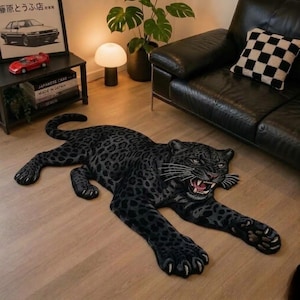 May include: A black panther-shaped area rug featuring a detailed dark gray leopard spot pattern, a snarling expression with visible teeth, and extended paws with claws. The rug is positioned on a light-colored wooden floor.
