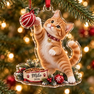 May include: A clear acrylic Christmas ornament shaped like a cat, featuring an orange tabby kitten wearing a red collar with a bell, reaching for a red and white yarn ball. Below, a food bowl is labeled with the name Milo and the year 2026.