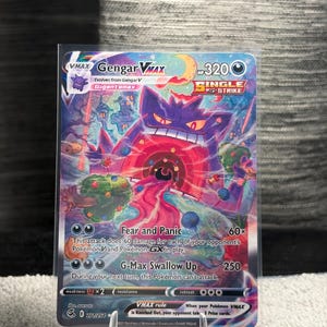 Puede incluir: Una carta coleccionable Gengar VMAX que muestra una gran criatura morada similar a un fantasma con la boca abierta, ojos rojos brillantes y un fondo oscuro con árboles coloridos y una luna creciente. La carta muestra 320 HP y texto.