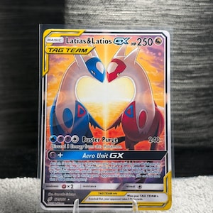 Puede incluir: Una carta coleccionable Latias & Latios GX Tag Team que muestra a los dos Pokémon legendarios frente a frente con un fondo de atardecer. La carta muestra 250 HP, los ataques Buster Purge y Aero Unit GX, y el texto 170/181 en la esquina.