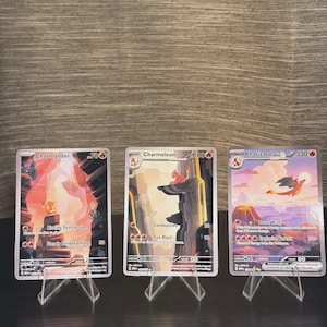 May include: Three Pokémon trading cards displayed on clear plastic stands. The cards feature Charmander, Charmeleon, and Charizard ex, each showcasing vibrant fire-themed illustrations, attack names like Blazing Destruction, Combustion, and Explosive Vortex, and their respective HP values of 70, 100, and 330.