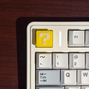 May include: A mechanical keyboard featuring a bright yellow custom keycap with a white question mark symbol in the top left corner. The keyboard has a white frame and light gray keys with black lettering, including standard keys like Tab and Caps Lock.