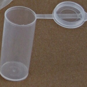 May include: A clear plastic vial with a hinged lid. The vial is cylindrical and has a smooth surface. The lid is attached to the vial with a small hinge.