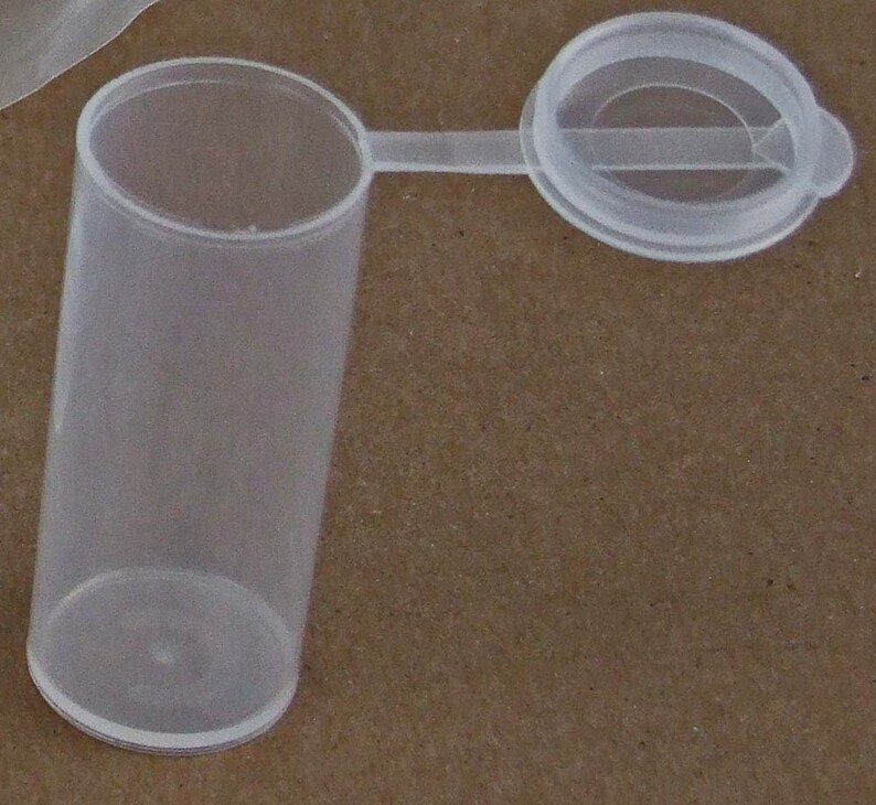 May include: A clear plastic vial with a hinged lid. The vial is cylindrical and has a smooth surface. The lid is attached to the vial with a small hinge.
