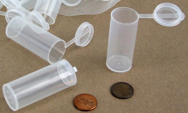 May include: A group of clear plastic vials with hinged lids. The vials are approximately 2.5 cm in diameter and 5 cm tall. The vials are sitting on a brown surface.