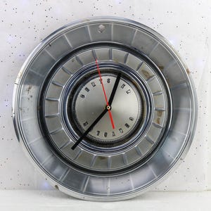 May include: A vintage chrome hubcap repurposed as a wall clock. The clock face features black hands and red hour markers. The clock is mounted on a white wall.
