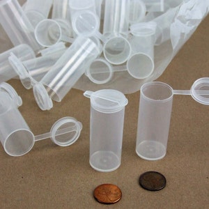 May include: A group of clear plastic containers with hinged lids. The containers are small and cylindrical, and they are arranged in a pile on a brown surface. Two of the containers are lying on their sides, with their lids open. The containers are about 2 inches tall and 1 inch in diameter.