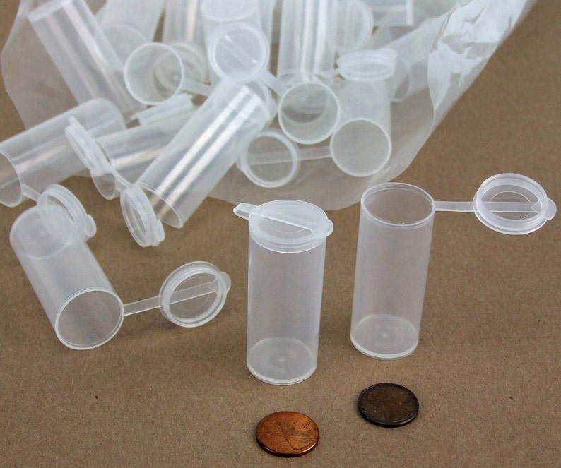 May include: A group of clear plastic containers with hinged lids. The containers are small and cylindrical, and they are arranged in a pile on a brown surface. Two of the containers are lying on their sides, with their lids open. The containers are approximately 5 cm tall and 2.5 cm in diameter.