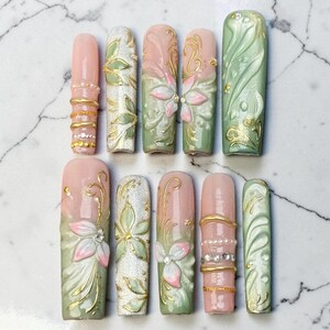 Op de afbeelding: Een set van tien kunstnagels met een mix van zachtroze en saliegroene tinten. De ontwerpen bevatten verhoogde 3D-bloemmotieven, gouden metallic accenten, glinsterende zilveren glittertexturen en kleine strass-steentjes in horizontale banden.
