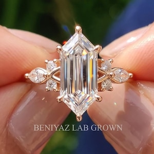 May include: A gold ring featuring a central elongated hexagonal cut stone flanked by smaller round and pear-shaped stones on each side. The band is thin and metallic. The text BENIYAZ LAB GROWN is printed in white at the base of the ring setting.