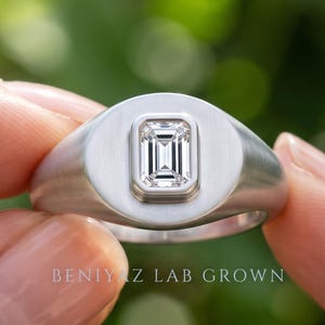 Silver tone emerald cut diamond signet ring held between fingers, showing the broad face and minimalist design.