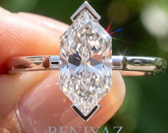 Puede incluir: Un anillo de tono plateado con una piedra central de corte marquesa sujeta en un engaste en forma de V. La banda es lisa y metálica, con pequeños detalles rectangulares a ambos lados de la piedra. El texto BENIYAZ LAB GROWN es visible.