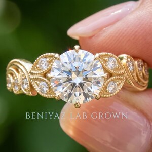 May include: A gold ring featuring a large round brilliant-cut center stone surrounded by smaller round stones set in a leaf-like pattern along the band. The text BENIYAZ LAB GROWN is visible below the ring. The metal has a polished yellow finish.