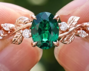 Puede incluir: Un anillo con una piedra preciosa verde de corte ovalado en una banda de metal color rosa. La banda presenta un diseño de enredadera con hojas esculpidas y pequeñas piedras claras redondas y de corte marquesa. Texto BENIYAZ LAB GROWN.