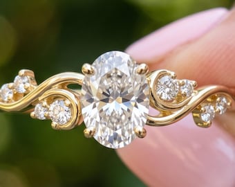 Puede incluir: Un anillo de oro con una piedra central de corte ovalado flanqueada por piedras redondas más pequeñas a lo largo de bandas metálicas curvas. El texto BENIYAZ LAB GROWN es visible en la parte inferior. Incluye varias piedras de acento.