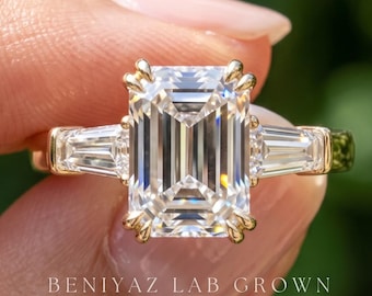 May include: A gold ring featuring a large emerald-cut centre stone flanked by two tapered baguette-cut side stones. The band is yellow gold, and the text BENIYAZ LAB GROWN is printed in white at the bottom of the image.
