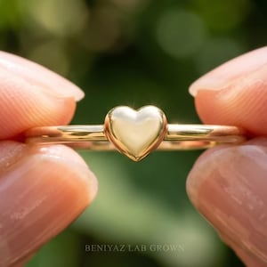 May include: A polished gold ring featuring a small, smooth heart-shaped centerpiece. The band is thin and circular, crafted from a reflective yellow metal. The text BENIYAZ LAB GROWN is printed in small, clear letters below the ring's center.
