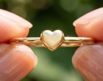 May include: A polished gold ring featuring a small, smooth heart-shaped centrepiece. The band is thin and circular, crafted from a reflective yellow metal. The text BENIYAZ LAB GROWN is printed in small, clear letters below the ring's centre.