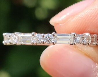 Puede incluir: Un anillo de banda en tono plateado que presenta un patrón alterno de piedras transparentes de corte baguette y piedras de corte brillante redondo, engastadas en una fila continua sobre la banda.