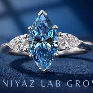 Blue marquise lab grown diamond engagement ring with pear cut side diamonds in a classic three stone setting on blue fabric.
