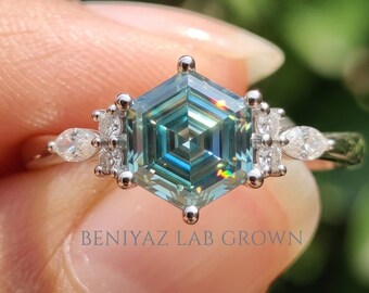 Puede incluir: Un anillo de tono plateado con una piedra preciosa central de color azul verdoso y corte hexagonal. La piedra central está flanqueada por piedras de acento más pequeñas, claras, de corte marquesa y redondo, montadas en una banda de metal. El texto BENIYAZ LAB GROWN es visible.