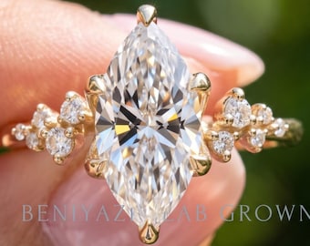 Puede incluir: Un anillo de oro con una gran piedra central de corte marquesa y piedras redondas más pequeñas a cada lado. La banda es de oro amarillo y el texto BENIYAZ LAB GROWN es visible en el centro de la montura del anillo.
