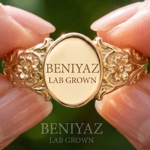 May include: A gold-toned ring featuring an oval-shaped center plate engraved with the text BENIYAZ LAB GROWN. The band displays intricate floral relief detailing on both sides of the central oval plate, showcasing a polished metallic finish.